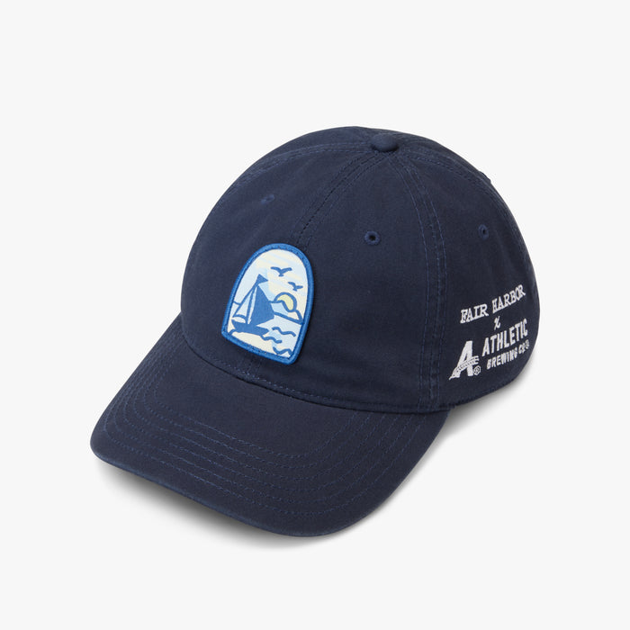 Navy blue baseball cap with a curved brim and embroidered graphics. The front features a blue-and-white patch showing a sailboat on the water with birds flying overhead and a sun near the horizon. The side of the hat includes white embroidered text reading ‘Fair Harbor x Athletic Brewing Co.’ on a light gray background.