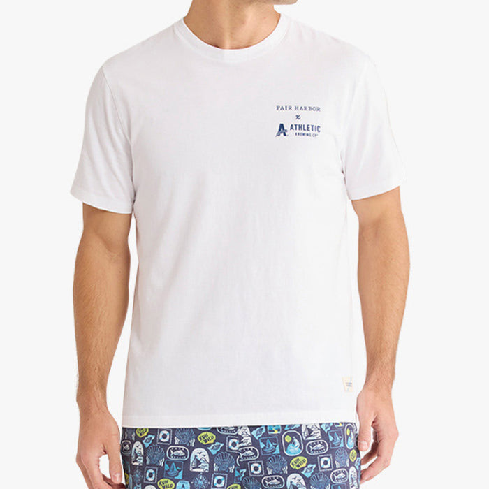 Front view of a person wearing a white short-sleeve T-shirt with a small blue ‘Fair Harbor x Athletic Brewing Co.’ logo printed on the left chest. The model is also wearing navy patterned swim shorts with nautical-inspired graphics.