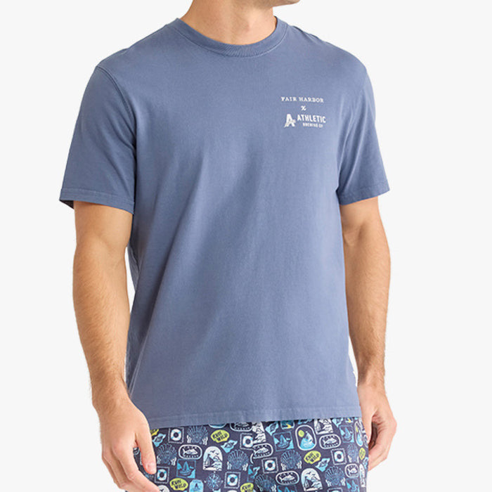 Front angled view of a person wearing a faded blue short-sleeve T-shirt with a small white ‘Fair Harbor x Athletic Brewing Co.’ logo printed on the left chest. The model is also wearing navy patterned swim shorts with coastal-themed graphics.