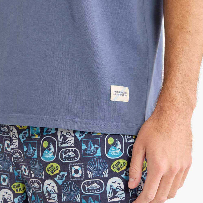 Close-up of the lower hem of a faded blue T-shirt featuring a small Fair Harbor fabric label. Patterned navy swim shorts with nautical-inspired graphics are partially visible below.