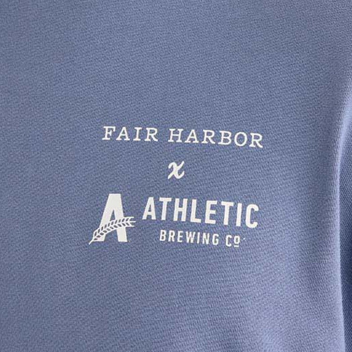 Close-up angled view of a faded blue short-sleeve T-shirt with a white ‘Fair Harbor x Athletic Brewing Co.’ logo printed on the left chest.