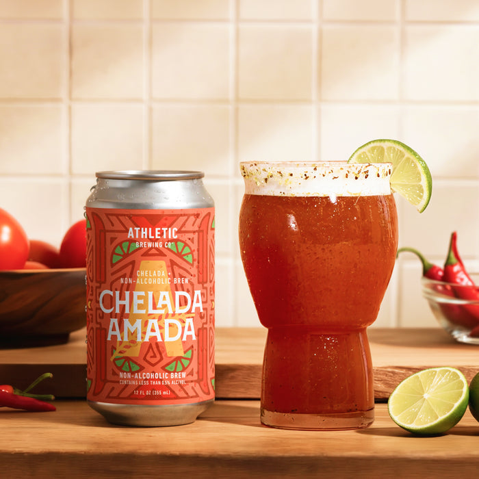 Can of Athletic Brewing Chelada Amada non-alcoholic brew beside a chilled glass of chelada on a wooden countertop. The reddish-orange drink is garnished with a lime wheel and served in a chili-seasoned rim glass. Fresh limes, tomatoes, and red chili peppers surround the scene against a warm tiled kitchen backdrop.