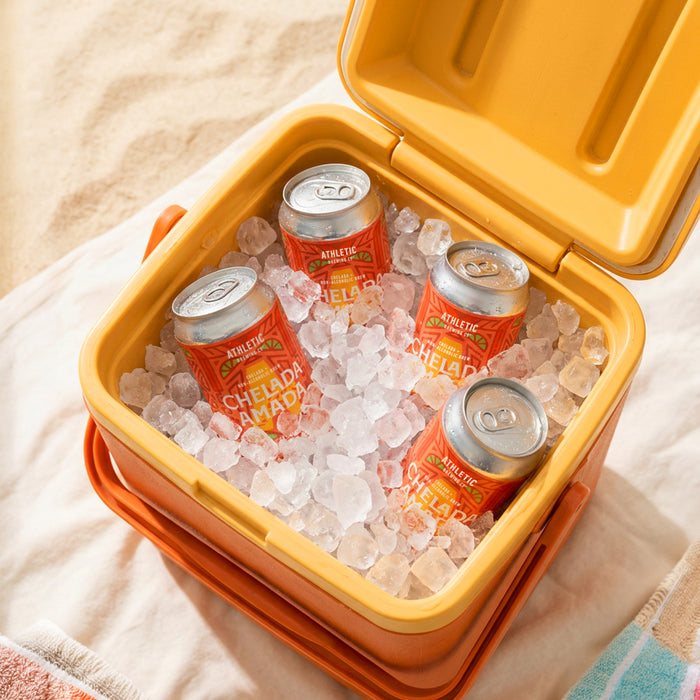 Four cans of Athletic Brewing Chelada Amada non-alcoholic brew nestled in crushed ice inside an open orange cooler. The cans are covered in condensation and arranged for easy grabbing, creating a refreshing outdoor scene on a light-colored blanket with a striped towel nearby.
