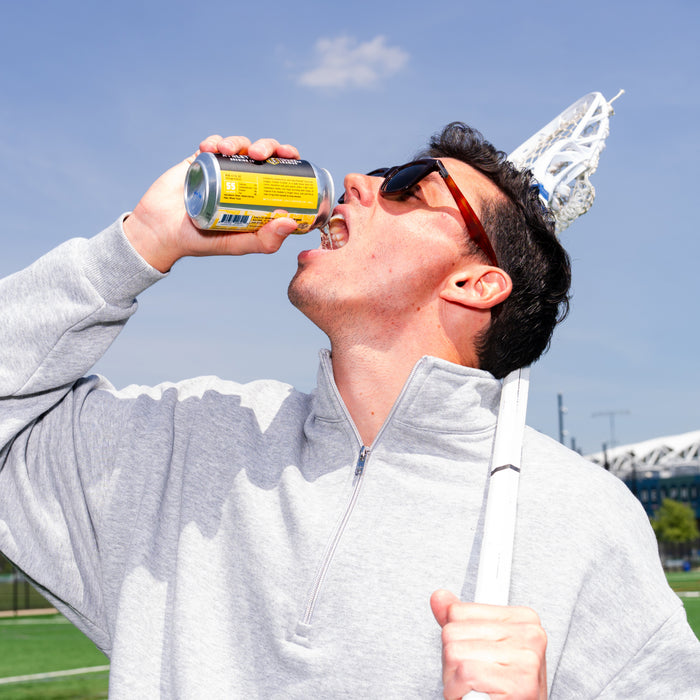 Person with grey quarter zip wearing sunglasses holding a lacrosse stick pouring a can of non alcoholic beer in their mouth