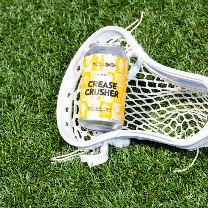 Can of Crease Crusher non alcoholic brew in a white lacrosse stick laying on artificial grass turf