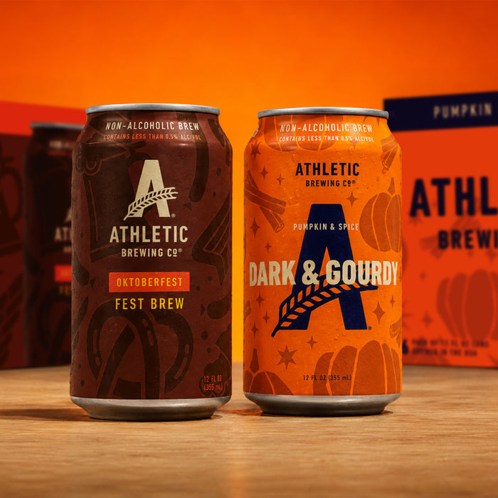 Athletic Brewing Oktoberfest Fest Brew and Dark & Gourdy Pumpkin & Spice non-alcoholic brew cans on a wooden surface with seasonal packaging in the background.