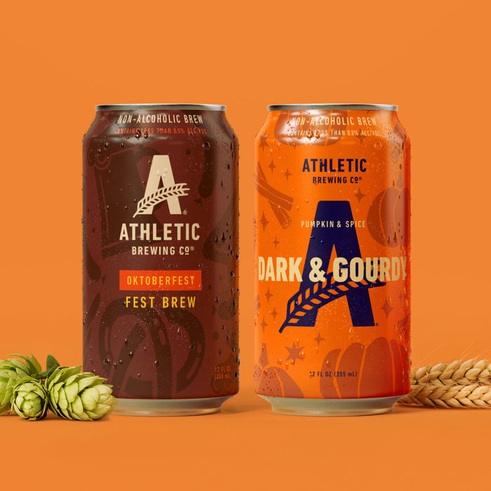 Athletic Brewing Oktoberfest Fest Brew and Dark & Gourdy Pumpkin & Spice non-alcoholic brew cans with condensation, displayed with hops and wheat on an orange background.