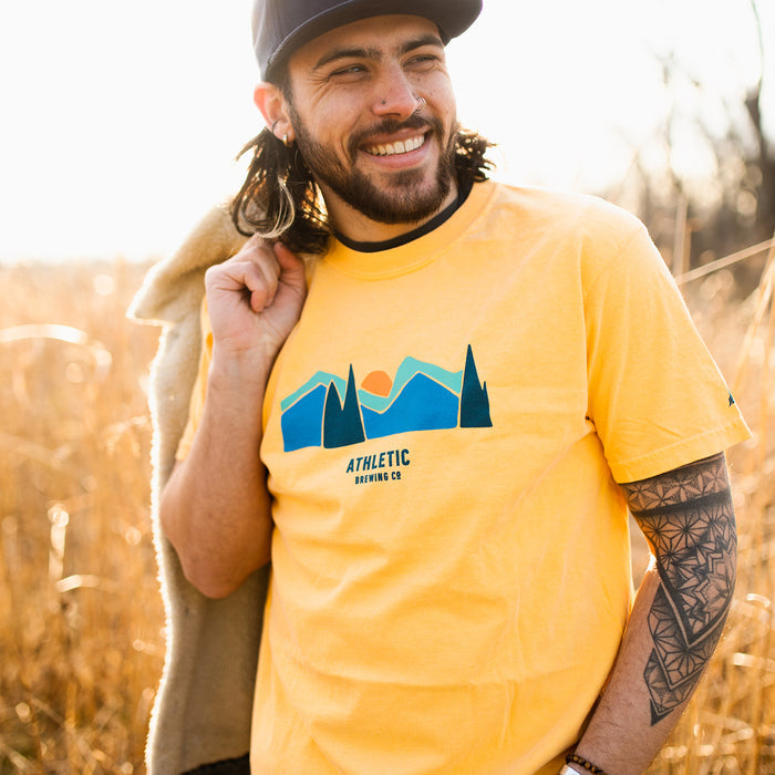 a man wearing A yellow Athletic brewing co flagship landscape t-shirt on a field