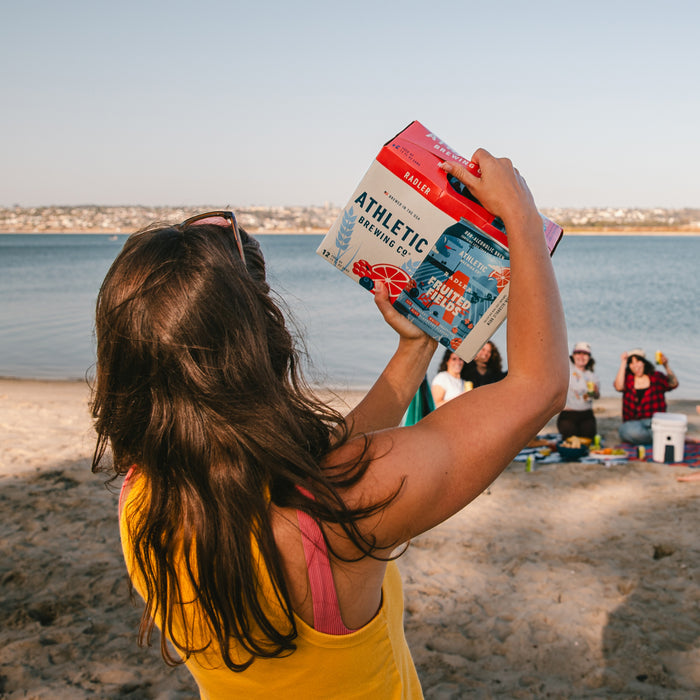 Person holding a 12 pack of Fruited Fields non alcoholic brew on the beach