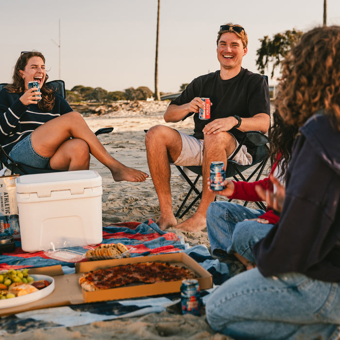 four people sitting on the beach holding cans of Fruited Fields Non alcoholic brew with a cooler and food on a blanket