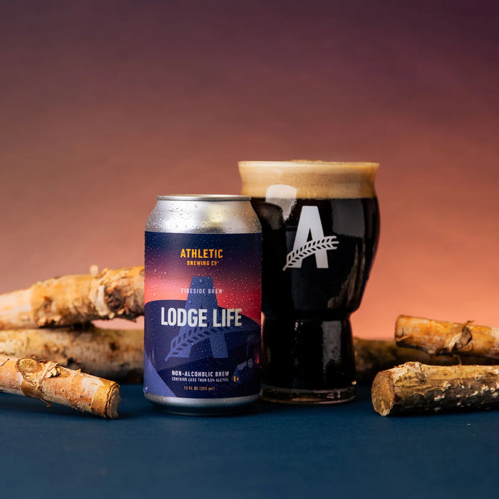 Athletic Brewing Co. Lodge Life non-alcoholic beer can and glass with wooden logs on a gradient background