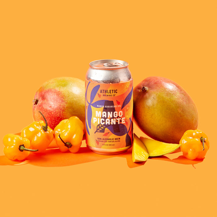 Athletic Brewing Mango Picante non-alcoholic mango habanero IPA can displayed on a bright orange surface and background, surrounded by whole mangoes, sliced mango pieces, and yellow habanero peppers. The colorful can features orange, yellow, and navy tropical graphics with habanero pepper illustrations. Water droplets on the chilled can and fresh fruit accents highlight the beer’s sweet-and-spicy flavor inspiration.
