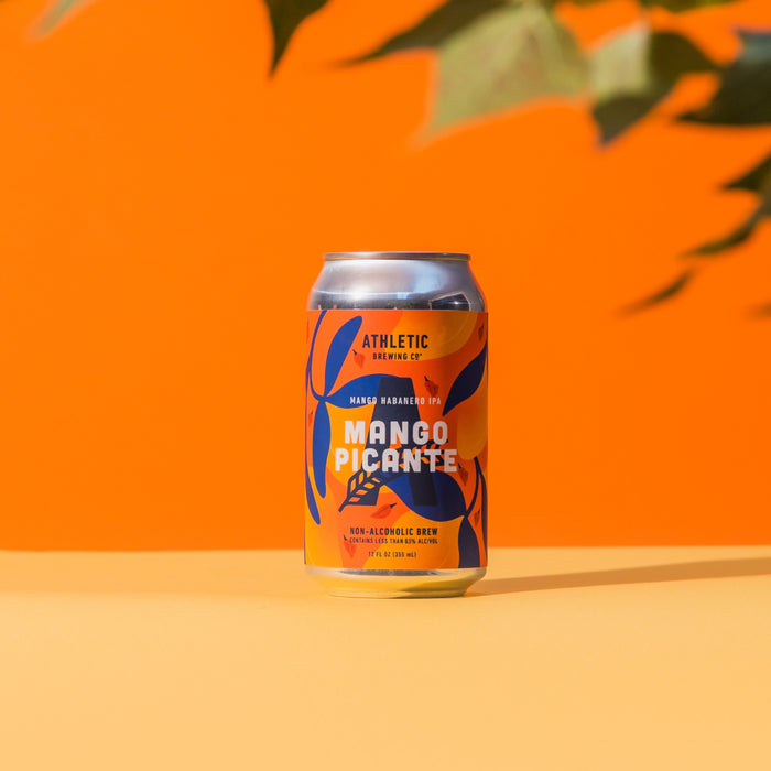 12 fl oz can of Athletic Brewing Mango Picante non-alcoholic brew standing on a light tan surface against a vibrant orange background. The can features a colorful orange, yellow, and navy design with tropical leaves and small habanero pepper illustrations. The label reads “Mango Picante” and “Mango Habanero IPA.” Soft shadows fall behind the can, while out-of-focus plant leaves frame the top corners of the image, creating a bright, tropical product scene.