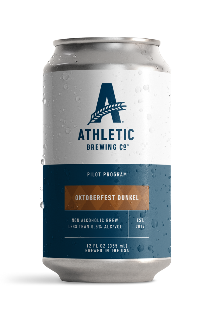 12 fl oz can of Athletic Brewing Oktoberfest Dunkel Pilot Program non-alcoholic brew on a white background. The can features Athletic Brewing's signature white and navy Pilot Program design with a warm brown label decorated with a subtle Bavarian diamond pattern. Condensation droplets cover the can, highlighting the beer's cold, refreshing presentation.