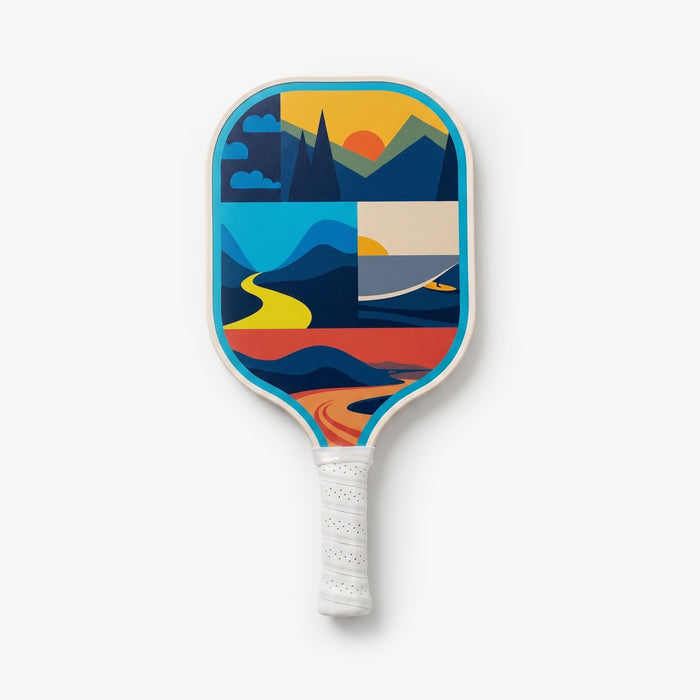 Back view of a pickleball paddle featuring a colorful abstract landscape design with mountains, water, clouds, and sunset-inspired graphics in blue, yellow, orange, and red tones. The paddle has turquoise trim and a white handle wrap.