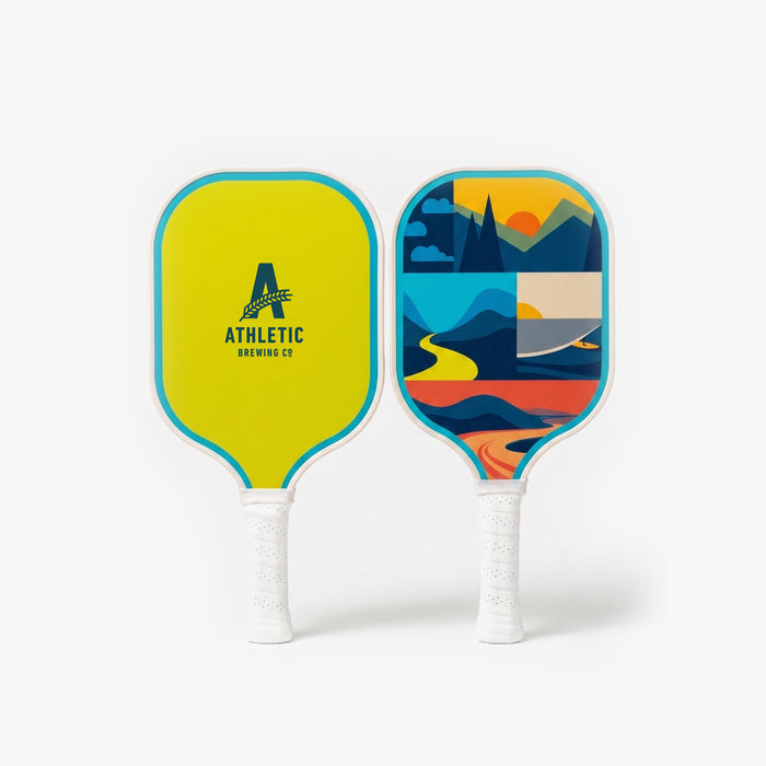 Two pickleball paddles standing side by side against a light gray background. One paddle features a neon yellow face with the Athletic Brewing Co. logo, while the other displays a colorful abstract landscape design in blue, yellow, orange, and red tones. Both paddles have turquoise trim and white handle wraps.