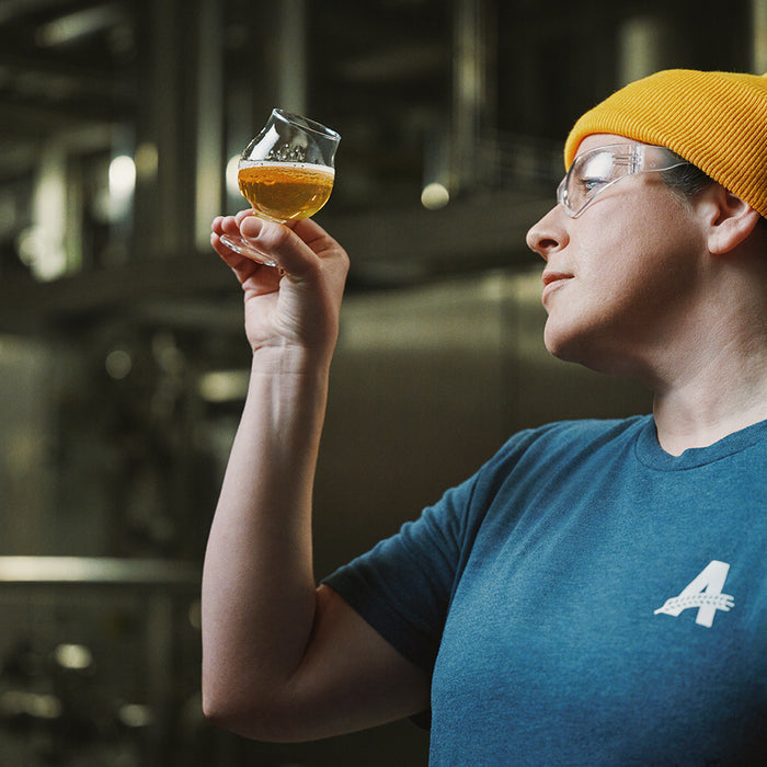 woman wearing a yellow beanie and safety glasses looking at a small glass of beer