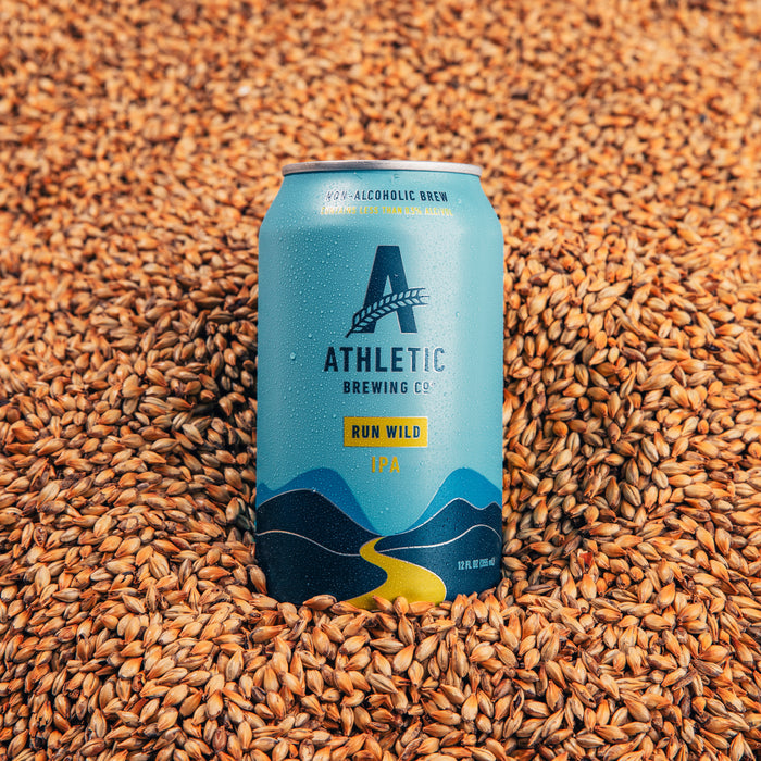 A cold can of Athletic Brewing Co. Run Wild IPA sits upright in a bed of malted barley grains. The light blue can features mountain artwork and visible condensation droplets, emphasizing refreshment and the beer’s non-alcoholic branding.